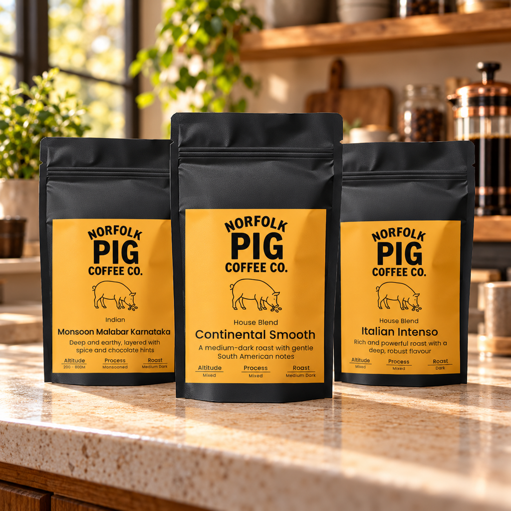 Three black coffee bags from Norfolk Pig Coffee Co. stand on a kitchen counter, with a blurred kitchen interior in the background.