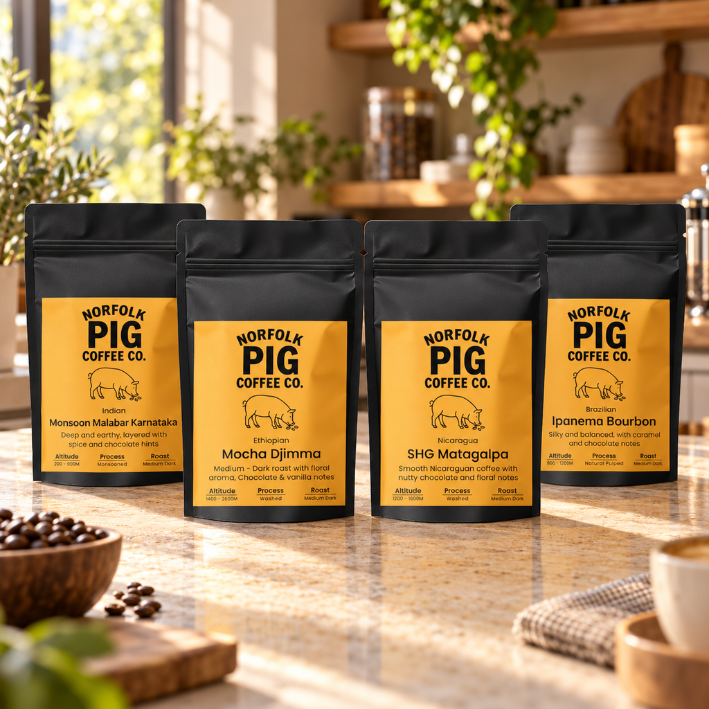 Four black stand-up coffee bags from Norfolk Pig Coffee Co. arranged on a kitchen counter with coffee beans, a bowl, and a cup in a warm, sunlit kitchen setting.