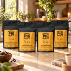 Four black stand-up coffee bags from Norfolk Pig Coffee Co. arranged on a kitchen counter with coffee beans, a bowl, and a cup in a warm, sunlit kitchen setting.
