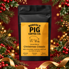 A black stand-up coffee bag with a large yellow label reading Norfolk Pig Coffee Co. Christmas Classic, centered against a festive holiday background with red fabric, holly, berries, and gold ribbon.