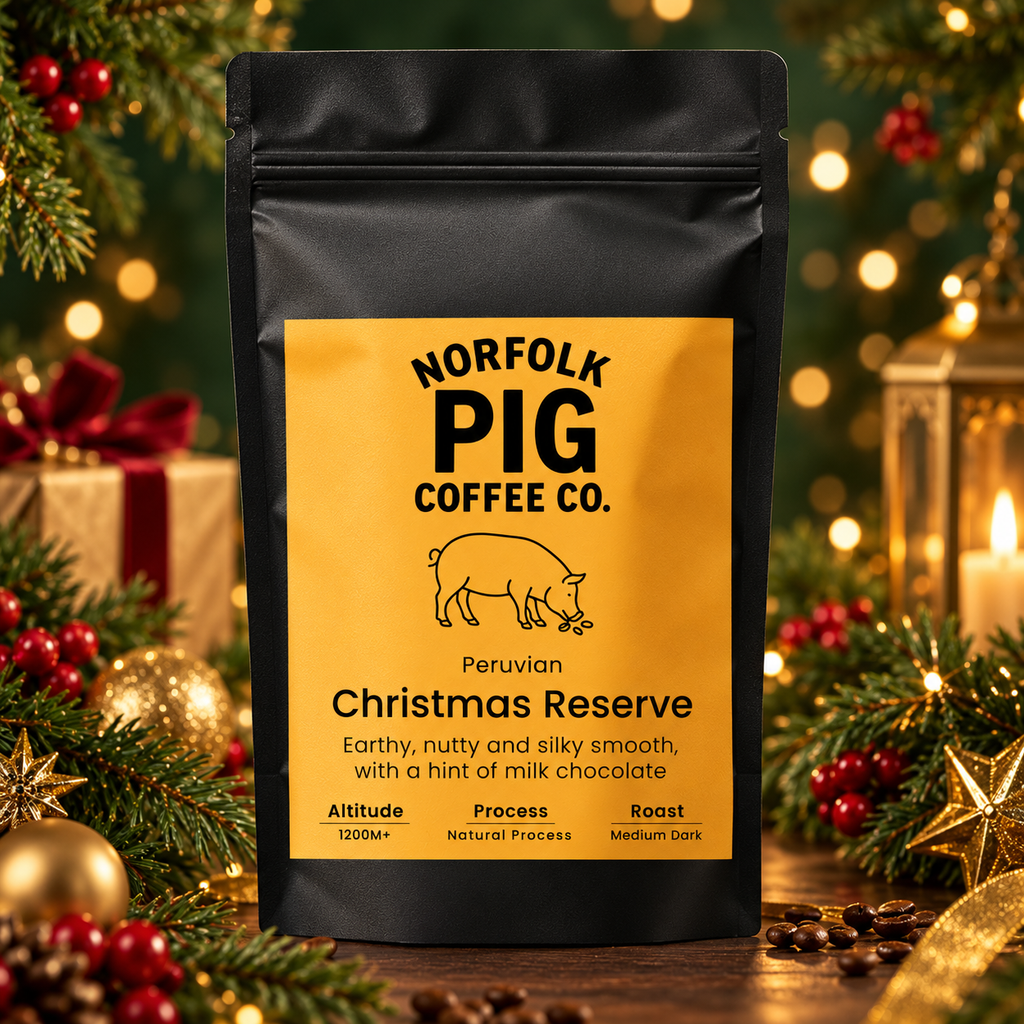 A black stand-up coffee bag labeled Norfolk Pig Coffee Co. Christmas Reserve stands on a tabletop surrounded by holiday greenery, ornaments, and warm blurred lights.