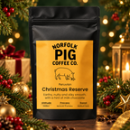 A black stand-up coffee bag labeled Norfolk Pig Coffee Co. Christmas Reserve stands on a tabletop surrounded by holiday greenery, ornaments, and warm blurred lights.