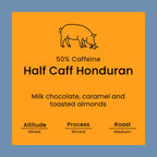 A square product label or packaging graphic with a bright orange center on a muted blue background. Black line art shows a pig at the top, with text reading '50% Caffeine', 'Half Caff Honduran', 'Milk chocolate, caramel and toasted almonds', and roast/processing notes at the bottom.