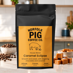 A black stand-up coffee bag labeled Norfolk Pig Coffee Co. Caramel Eclipse House Blend, standing on a kitchen counter with coffee beans and caramel cubes around it.