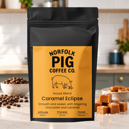 A black stand-up coffee bag labeled Norfolk Pig Coffee Co. Caramel Eclipse House Blend, standing on a kitchen counter with coffee beans and caramel cubes around it.