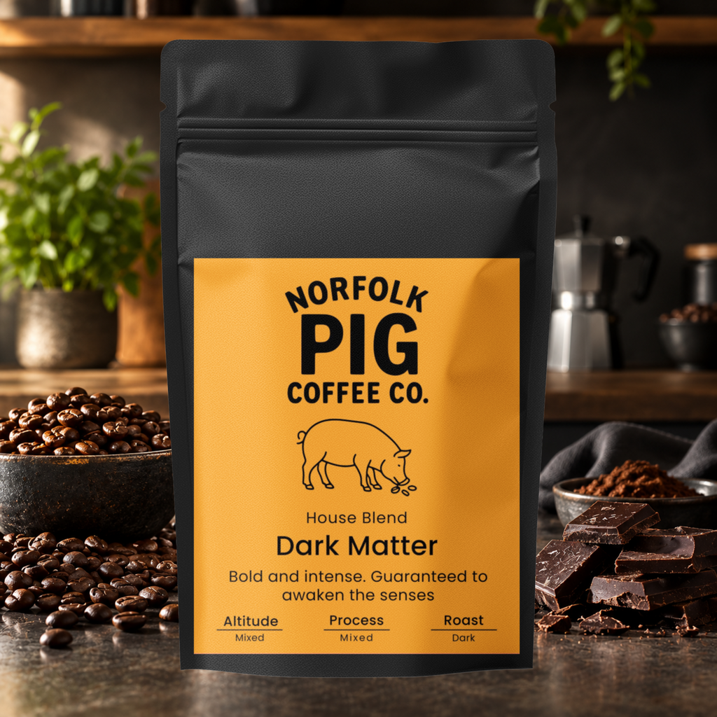 A black stand-up coffee bag labeled Norfolk Pig Coffee Co. Dark Matter House Blend, photographed on a rustic kitchen counter with coffee beans, chocolate, and a moka pot in the background.