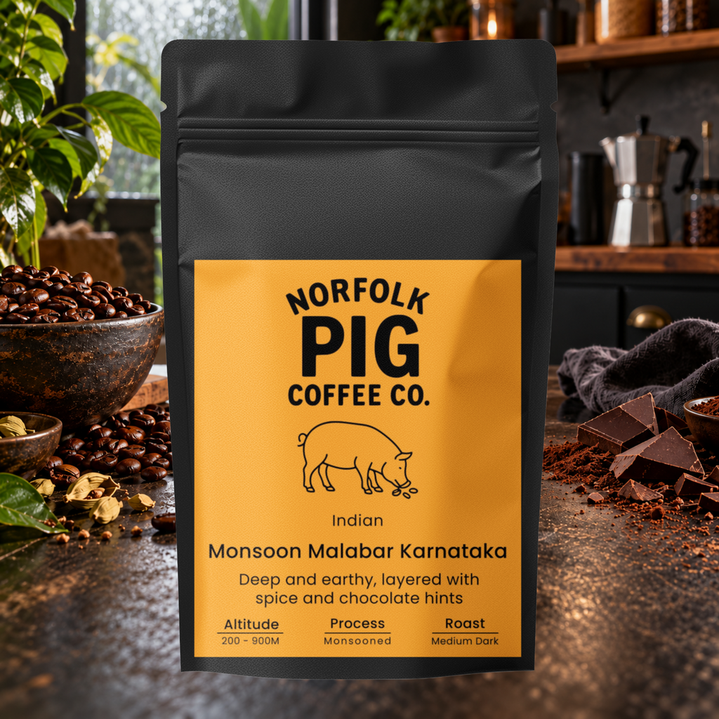 A black stand-up coffee bag labeled Norfolk Pig Coffee Co. Indian Monsoon Malabar Karnataka, standing on a countertop with coffee beans, chocolate, and kitchen props in a real indoor setting.