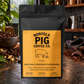 A black stand-up coffee bag labeled Norfolk Pig Coffee Co. Indian Monsoon Malabar Karnataka, standing on a countertop with coffee beans, chocolate, and kitchen props in a real indoor setting.