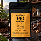 A black stand-up coffee bag labeled Norfolk Pig Coffee Co. Blue Mountain, placed in a kitchen-like setting with coffee beans, chocolate, and a moka pot in the background.