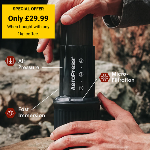 A person's hands holding a black AeroPress coffee maker against a rocky outdoor background, with promotional text and callouts highlighting air pressure, micro-filtration, and fast immersion.