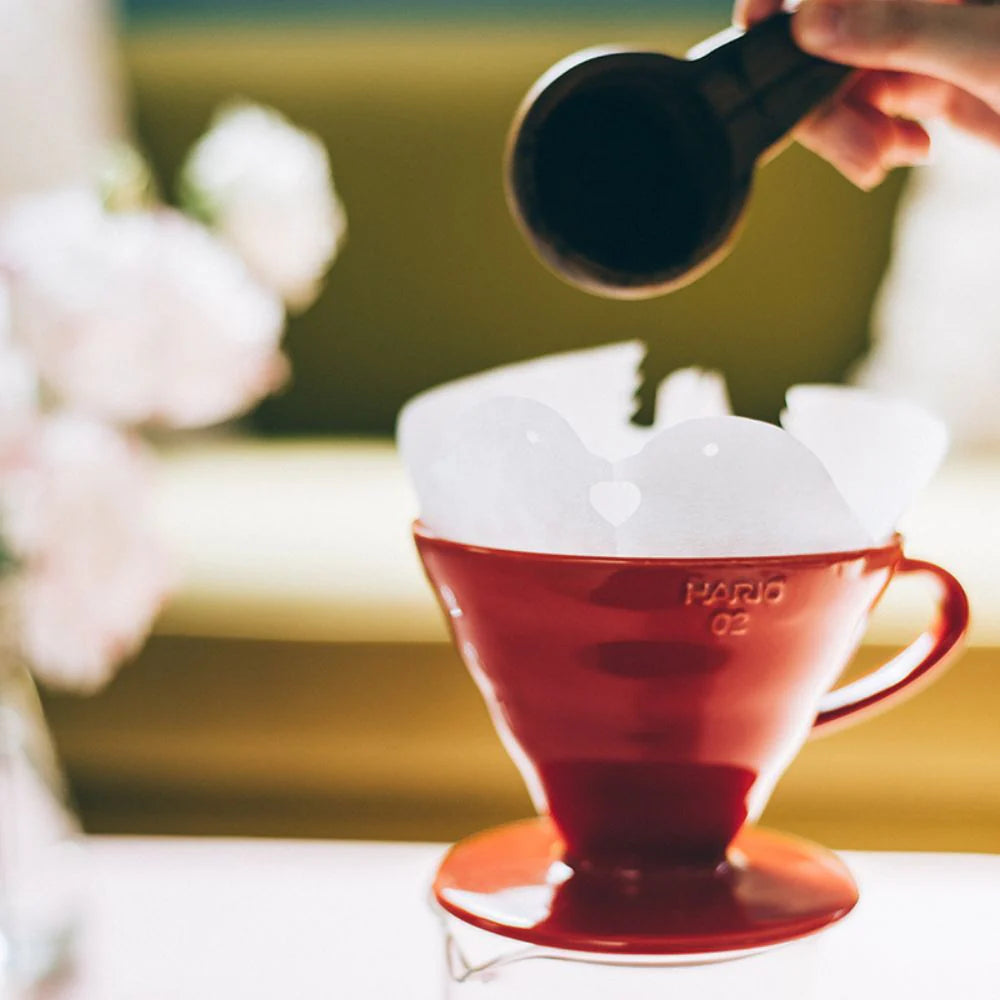 Red Hario coffee dripper with a hand pouring coffee into it, blurred background